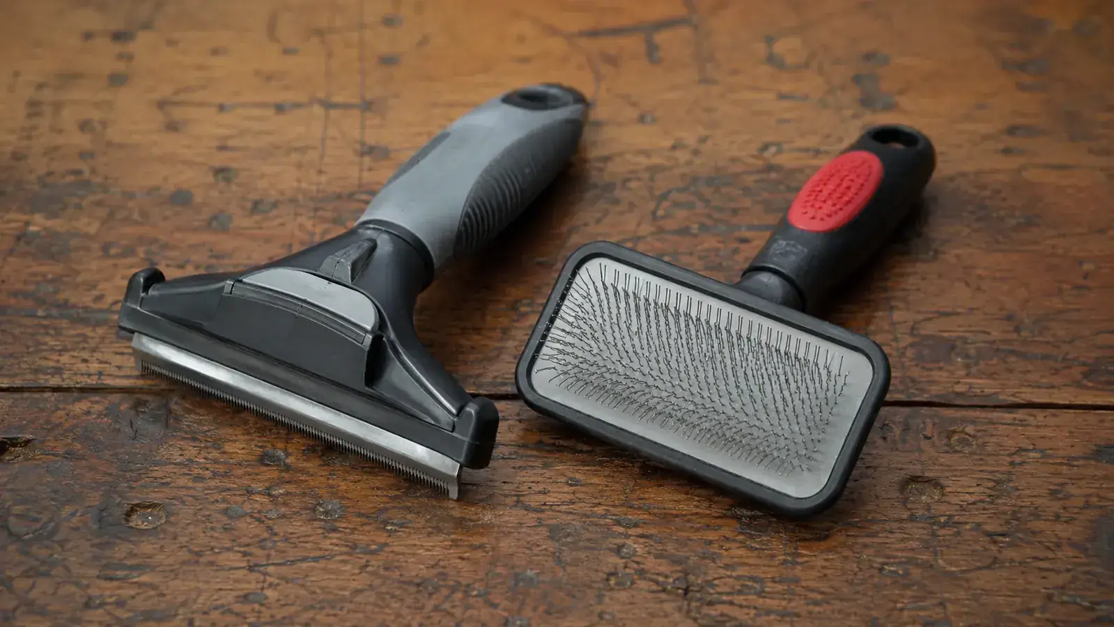 A deshedding tool and a slicker brush laid side by side on a worn wooden bench